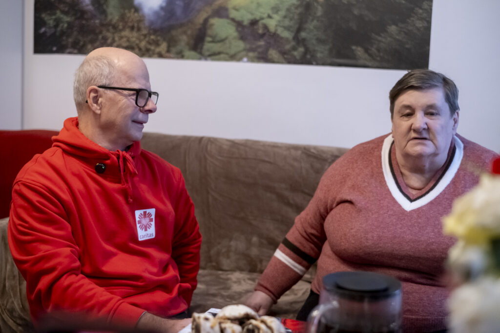 Wolontariusz w czerwonej bluzie Caritas i seniorka rozmawiają na kanapie przy stole z ciastem