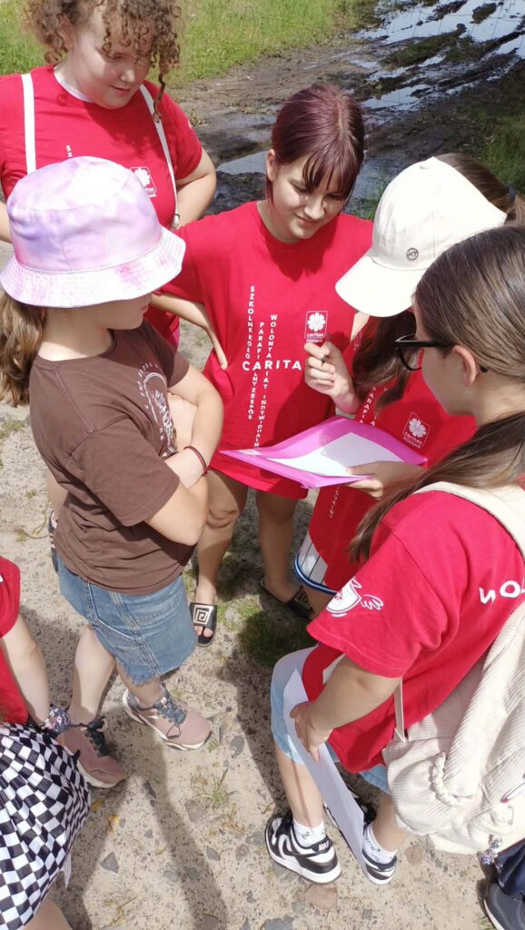 Wolontariuszki w koszulkach Caritas pochylają się nad różową teczką z zadaniem