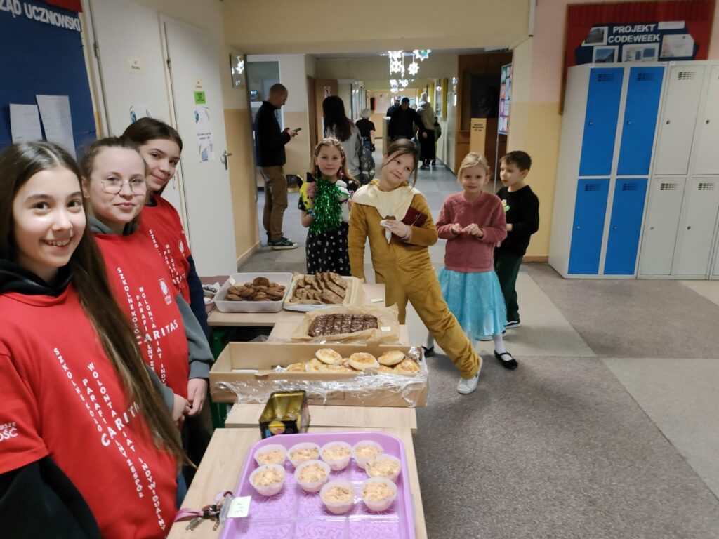 Wolontariuszki SKC przy stole z ciastami na szkolnym korytarzu, obok dzieci w przebraniach