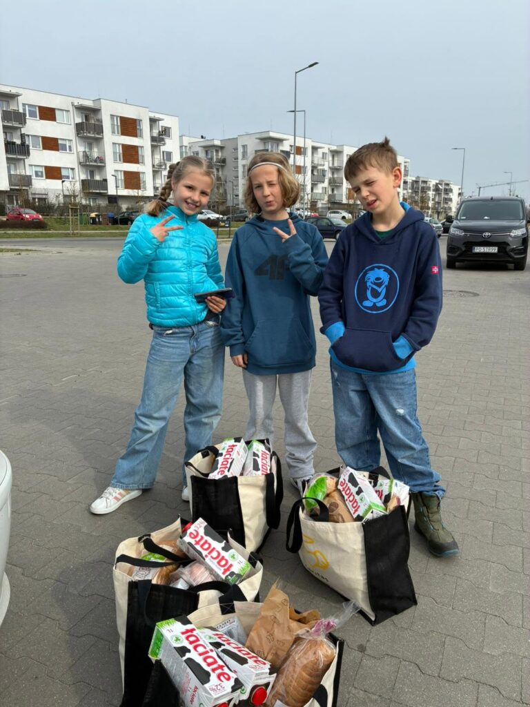 Troje dzieci z torbami wypełnionymi zakupami spożywczymi na parkingu osiedla