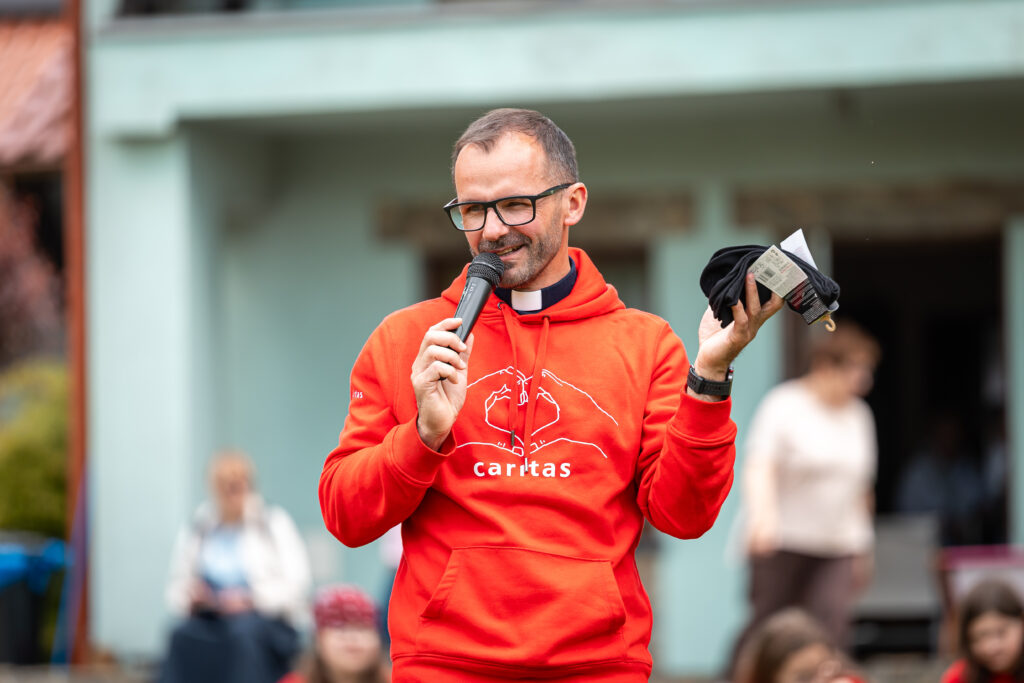 Pięcioro młodych wolontariuszy w koszulkach Caritas idzie po trawniku z jajkami na łyżkach