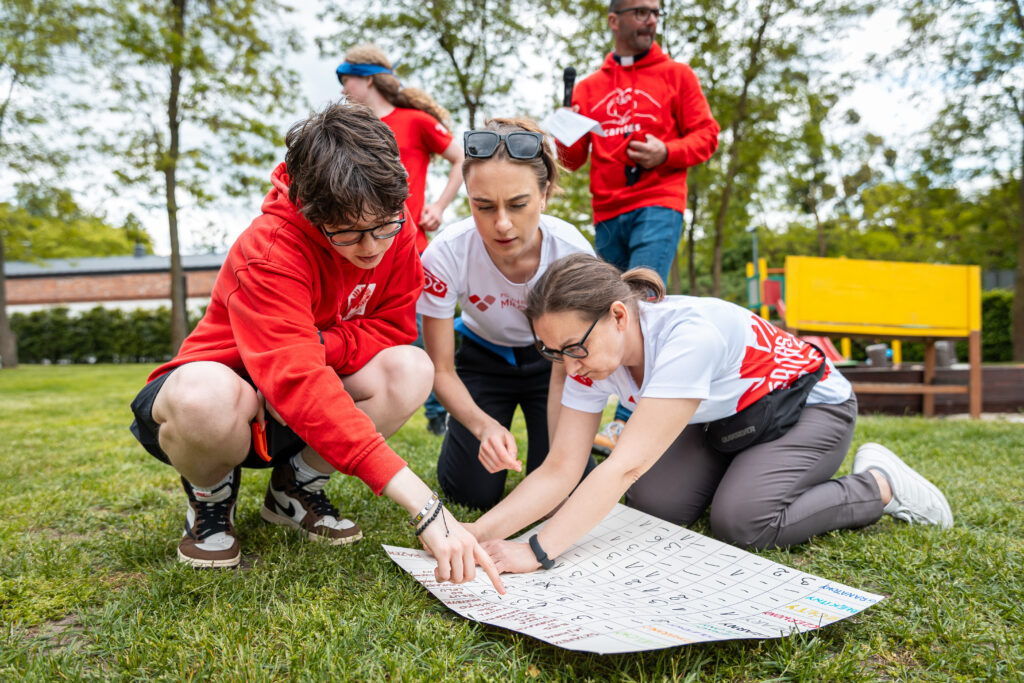 Chłopak i dwie wolontariuszki przy planszy z zadaniem rozłożonej na trawie